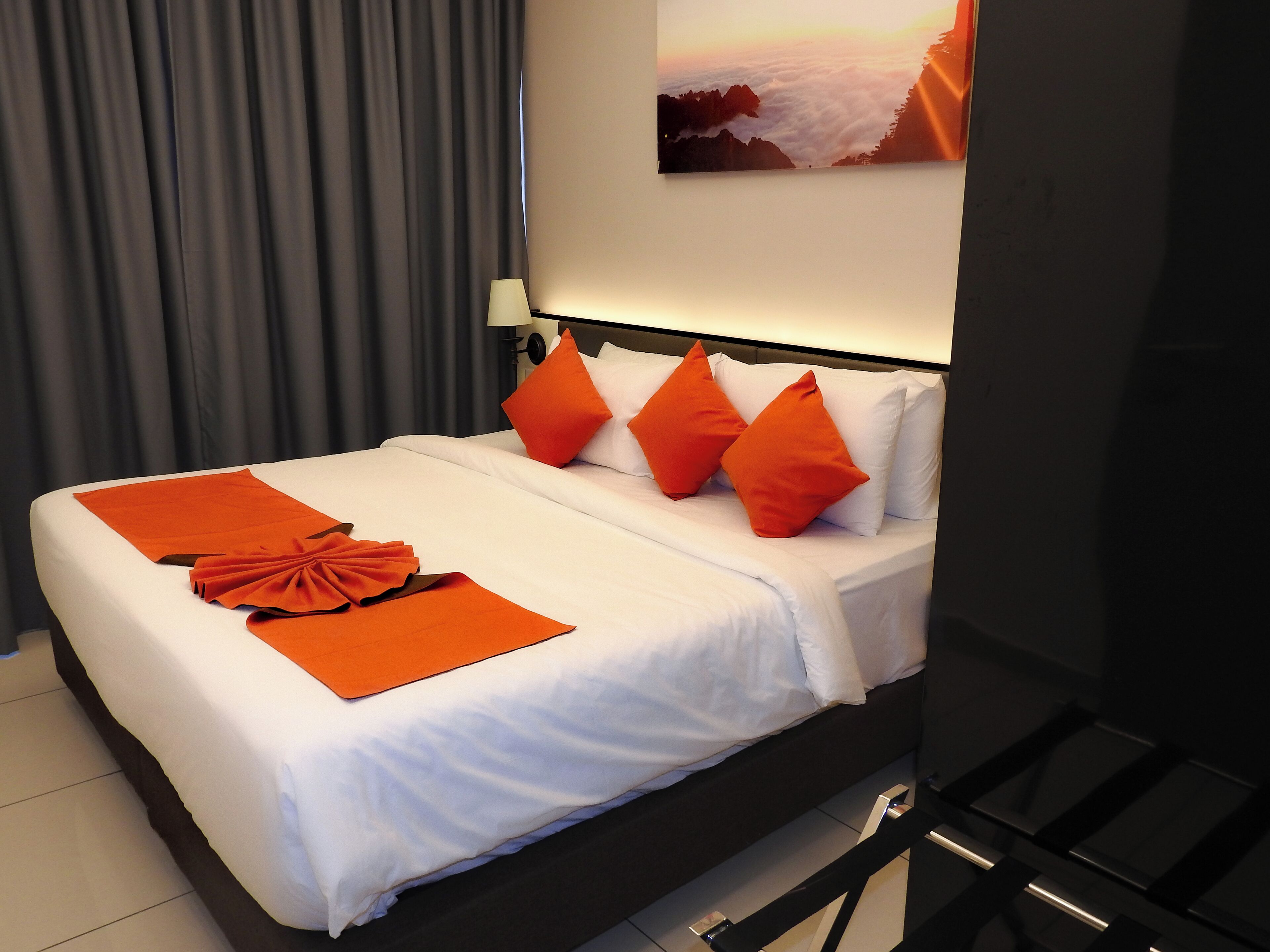 A neatly made bed with white bedding and orange pillows, a grey curtain, and a painting of a mountain landscape on the wall.