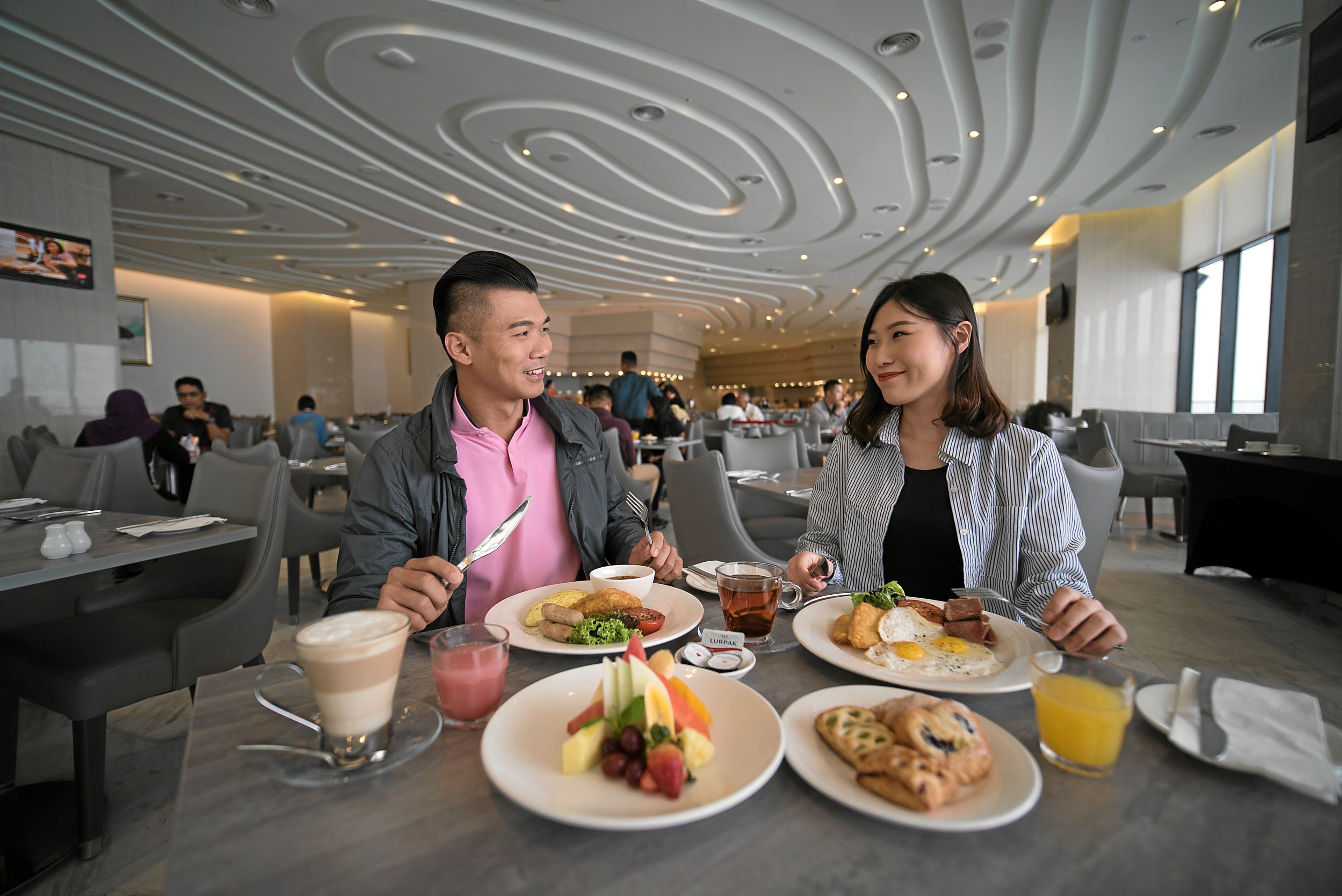 A modern dining area with a contemporary ceiling design, multiple dining tables, and a variety of breakfast dishes.