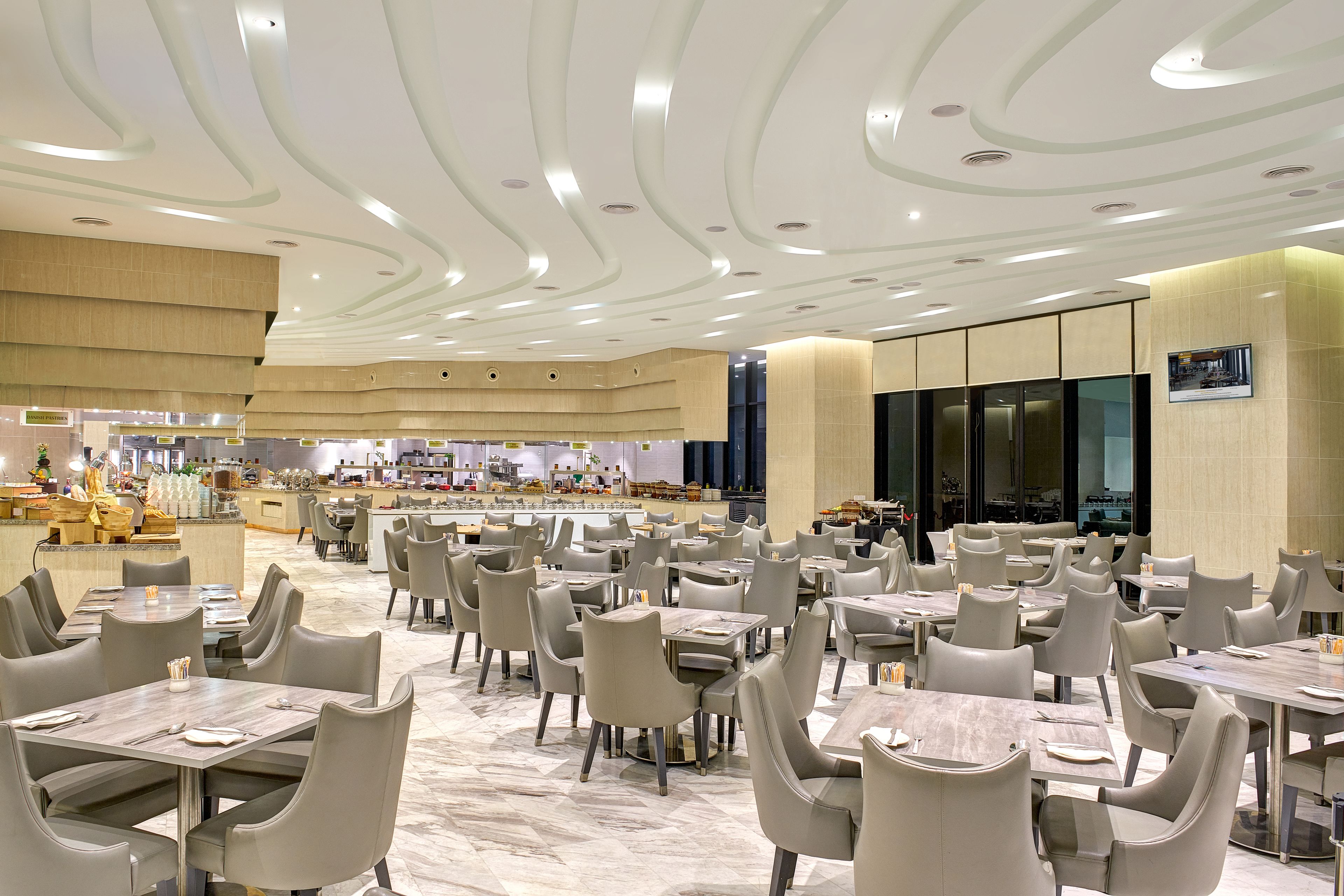 A spacious dining area with multiple tables and chairs, a modern ceiling design, and a visible kitchen area in the background.