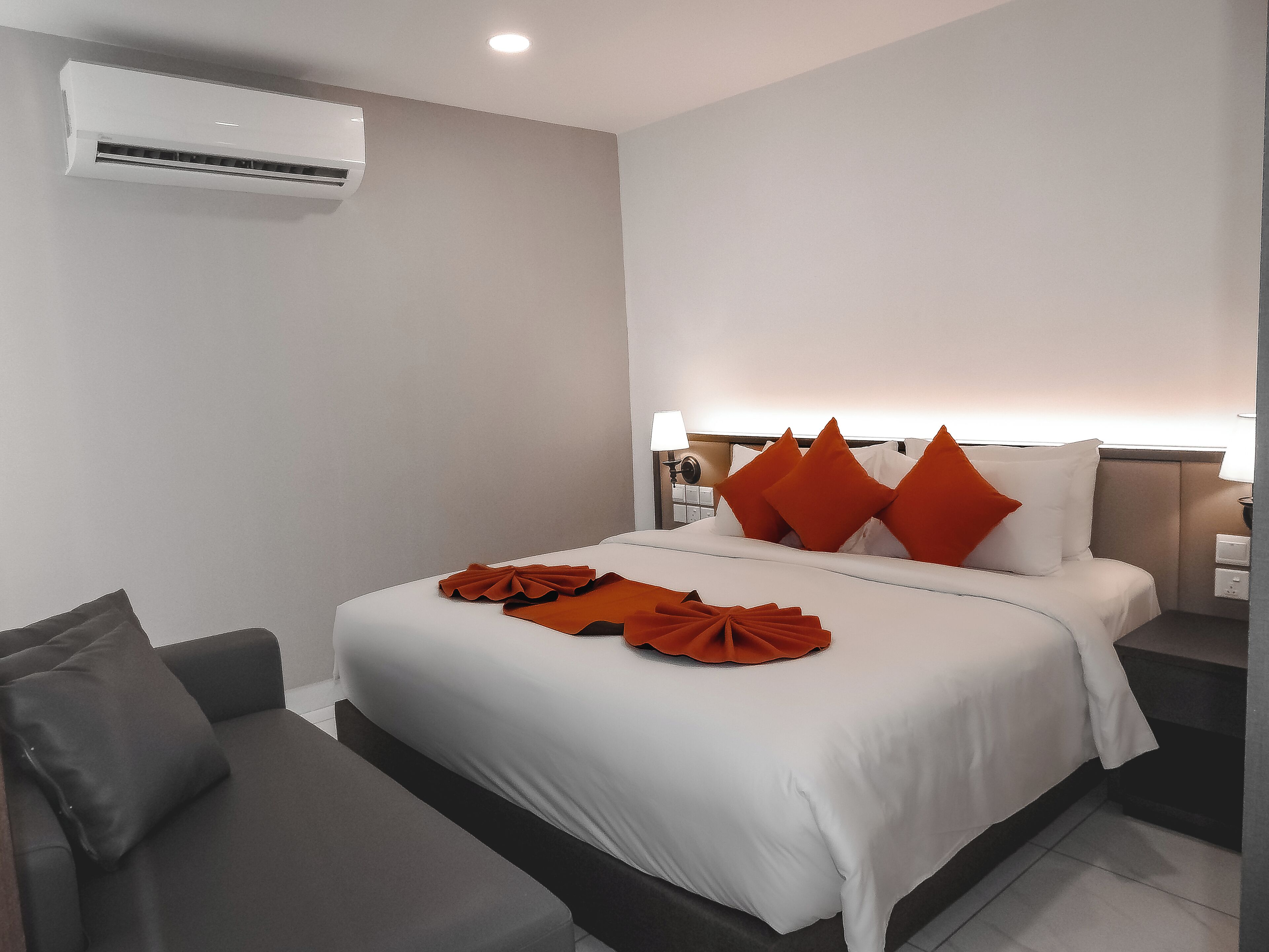 A hotel room with a bed, a bedside table, a nightstand, a lamp, and an air conditioning unit.