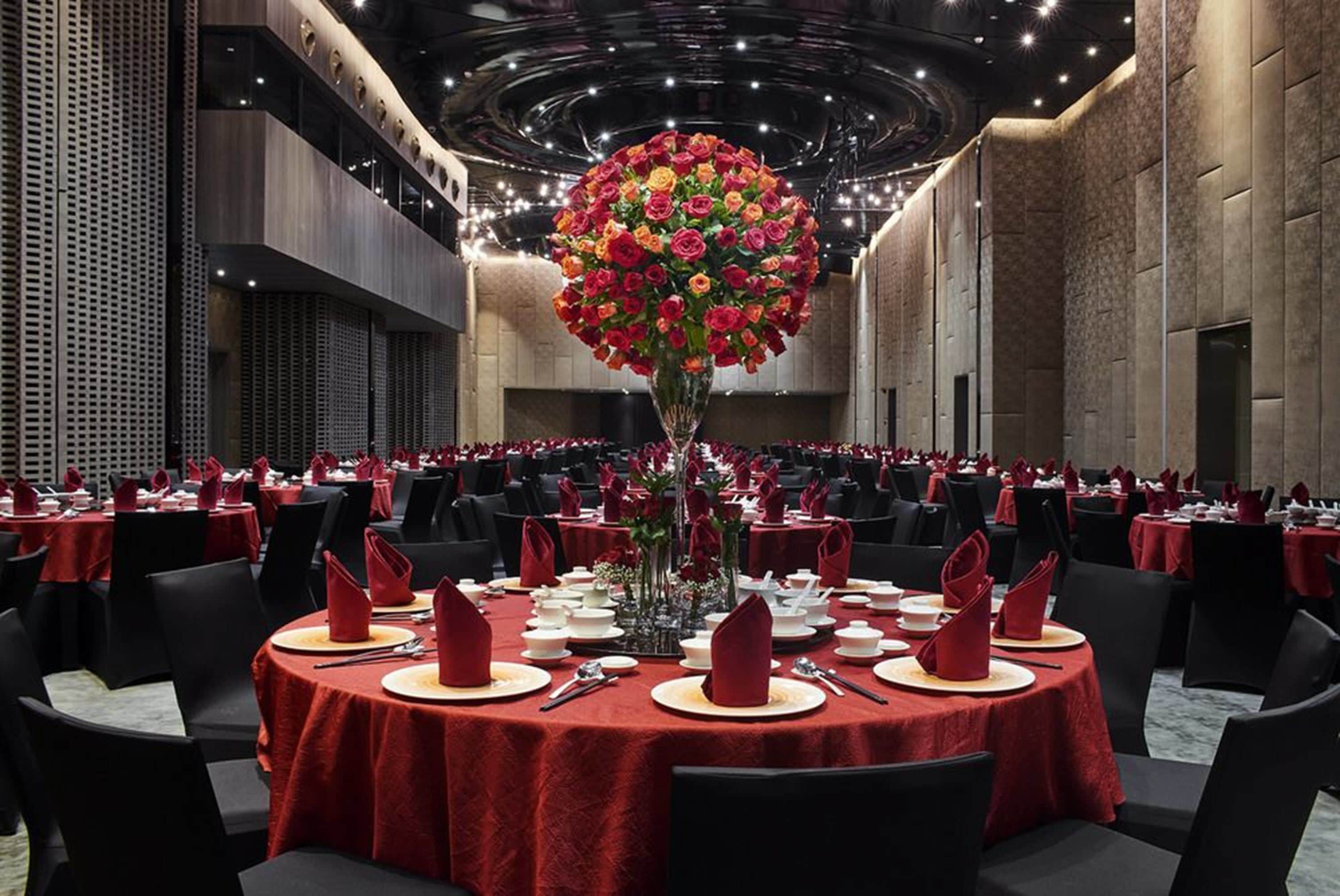 Un gran salón de banquetes con mesas redondas dispuestas para el servicio de comidas, con manteles rojos y un arreglo floral central.
