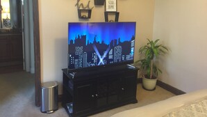 A flat-screen TV on a black stand, a framed picture with a cat, a clock, and a 'STAY HOME' sign on the wall.