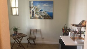 A small, cozy room with a dining area, a chair, a table, a potted plant, and a painting of a coastal scene.