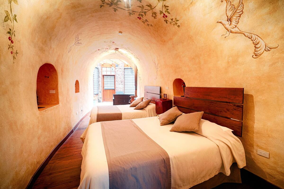 Une pièce confortable à l’atmosphère chaleureuse, avec des murs en arc, deux lits dotés de têtes de lit en bois et une fresque représentant un oiseau.