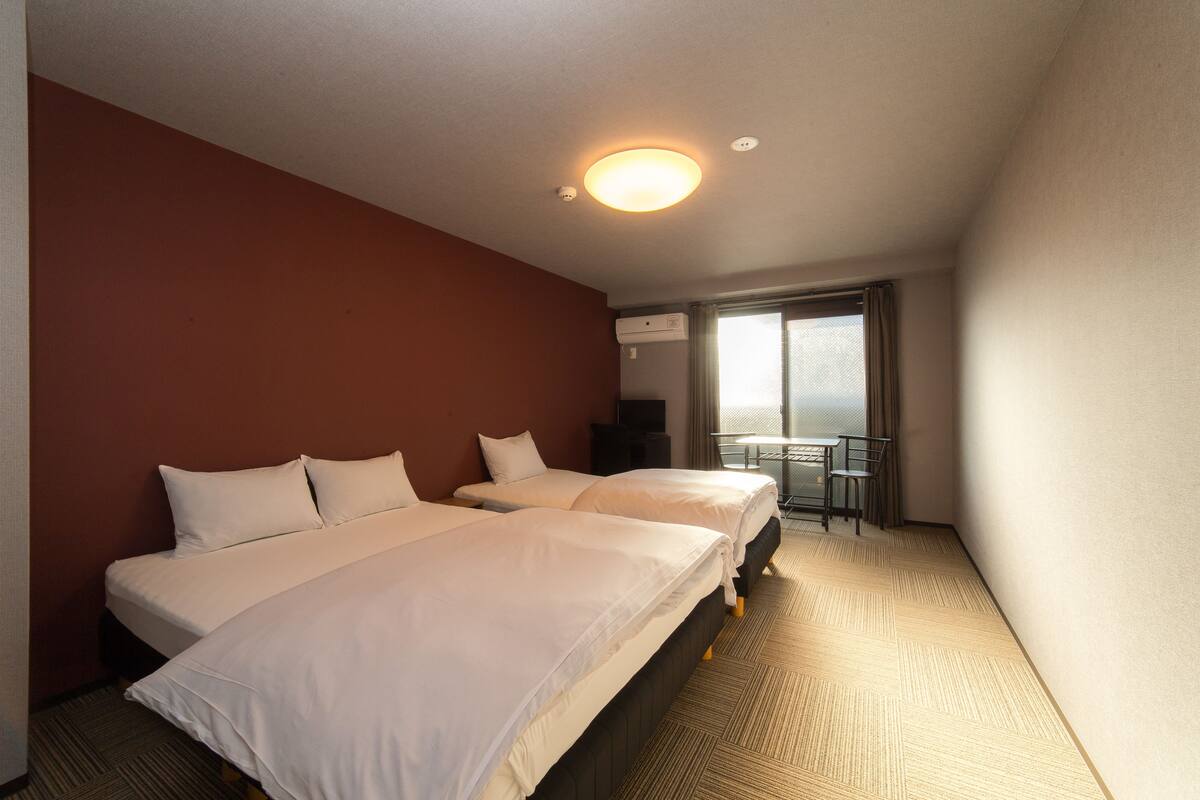 A hotel room with two beds, a small table, and a window with a view.