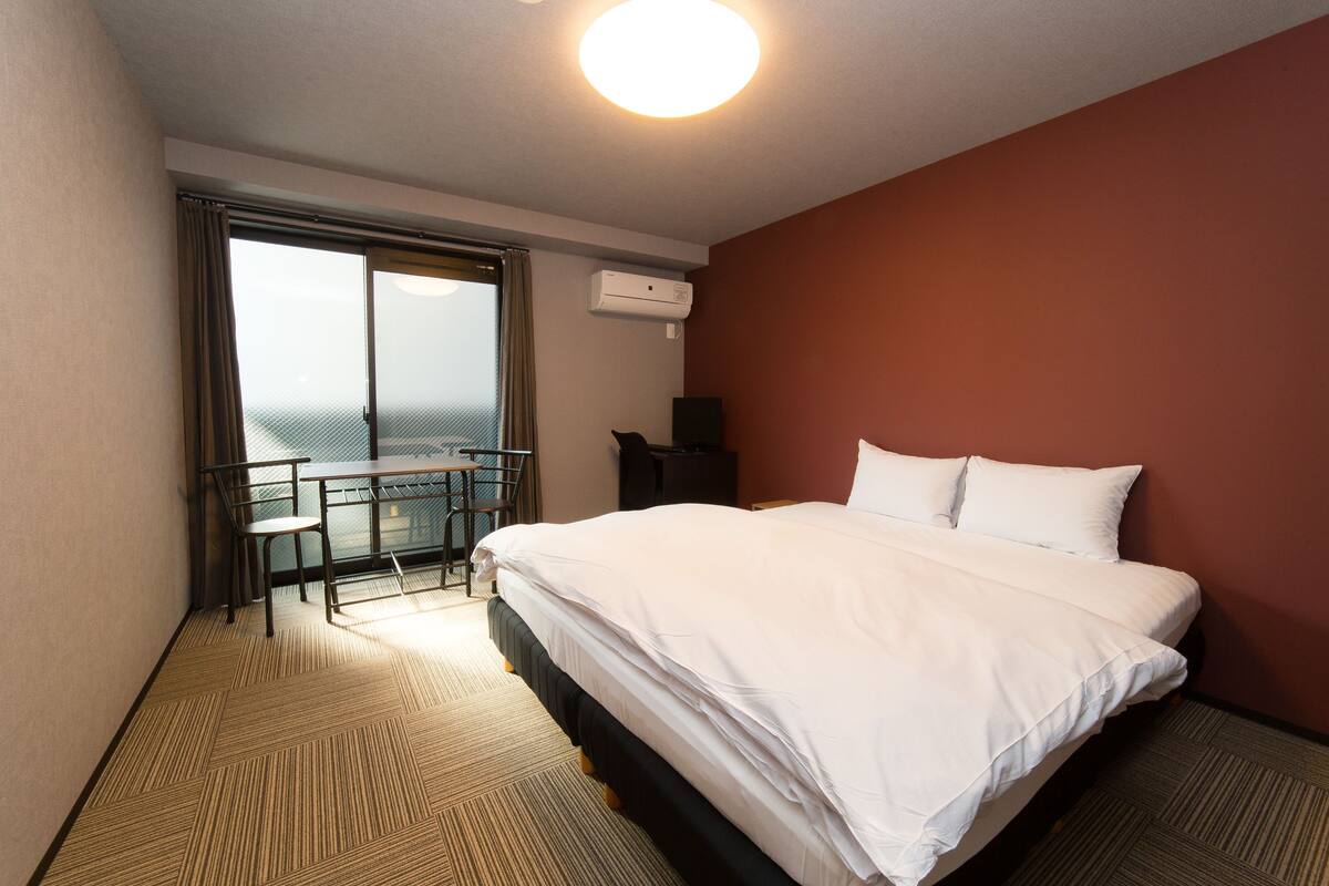 A hotel room with a large bed, a desk with a chair, a view through a window, and an air conditioning unit.