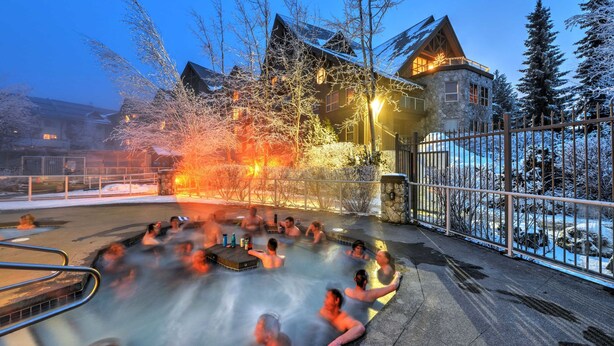 A hot tub area with people relaxing, surrounded by snow-covered trees and a stone building with a wooden roof. - Aspens #113 Prime Ski-in Ski-out Location! Pool, Hot tubs, BBQ, sleeps 4 (Whistler)
