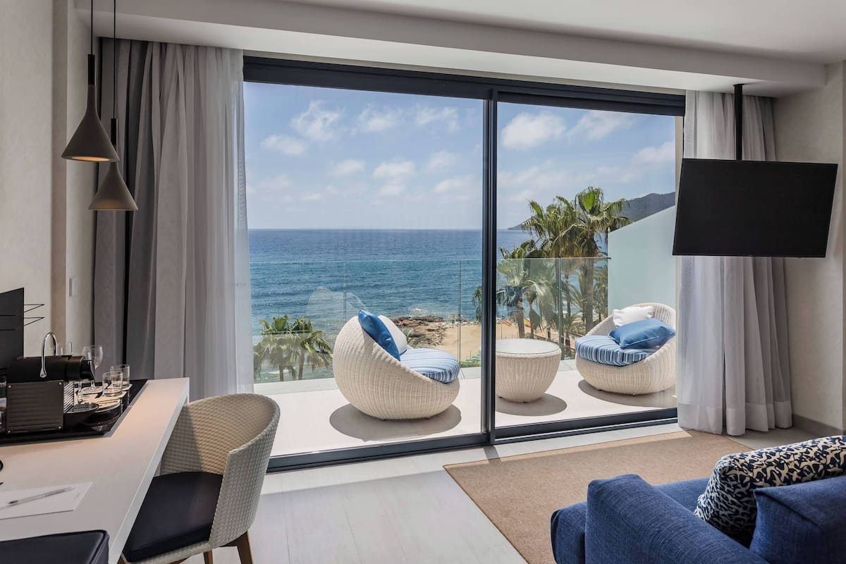 A modern room with a large sliding glass door leading to a balcony with a view of the ocean. There are two white wicker chairs with blue cushions, a white sofa, and a flat-screen TV mounted on the wall.