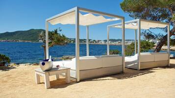 Un espace aménagé avec des sièges, situé en bord de plage, doté de parasols blancs et de meubles en osier, offrant une vue sur le paysage côtier.
