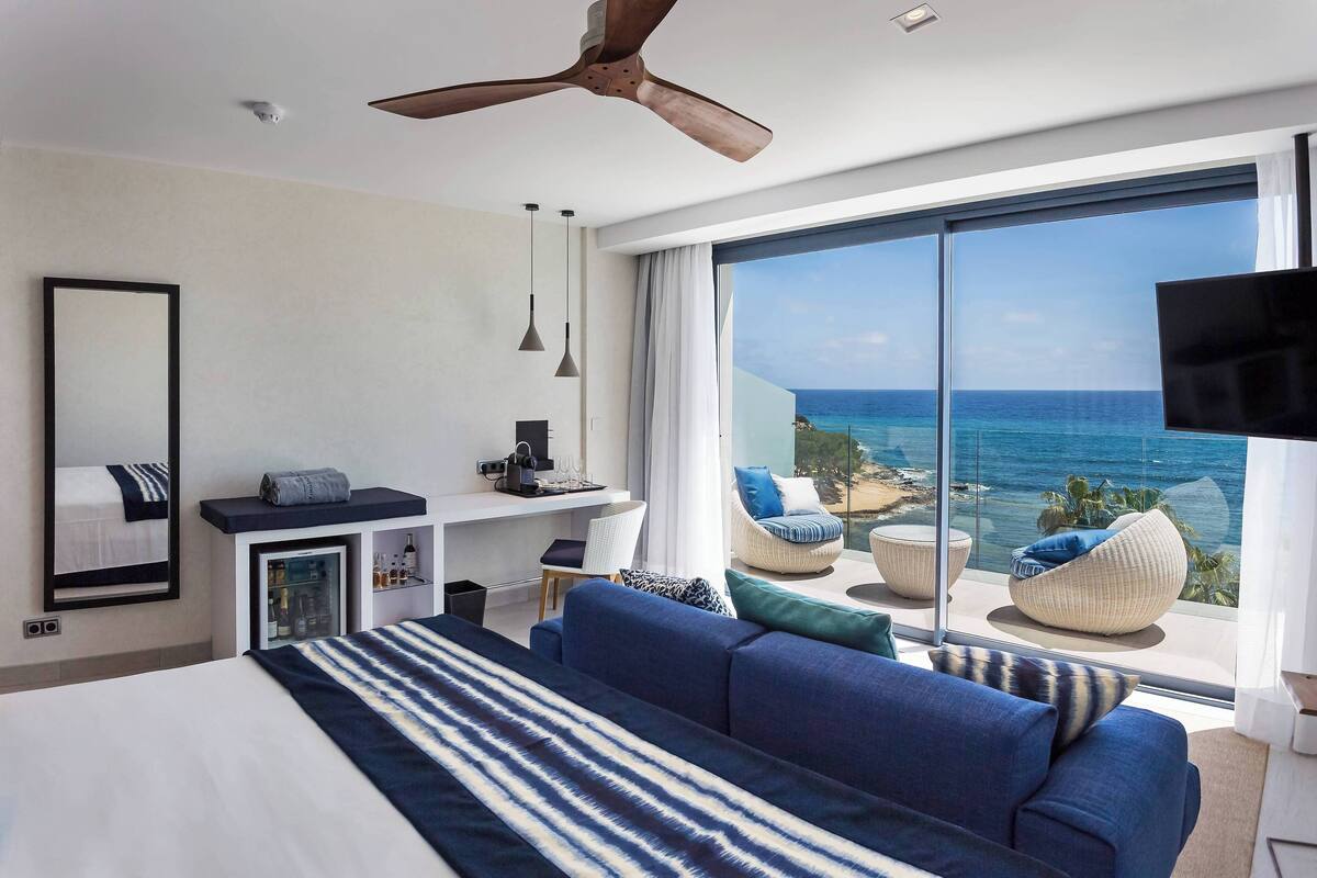 A modern hotel room with a large bed, a desk, a flat-screen TV, and a view of the ocean.