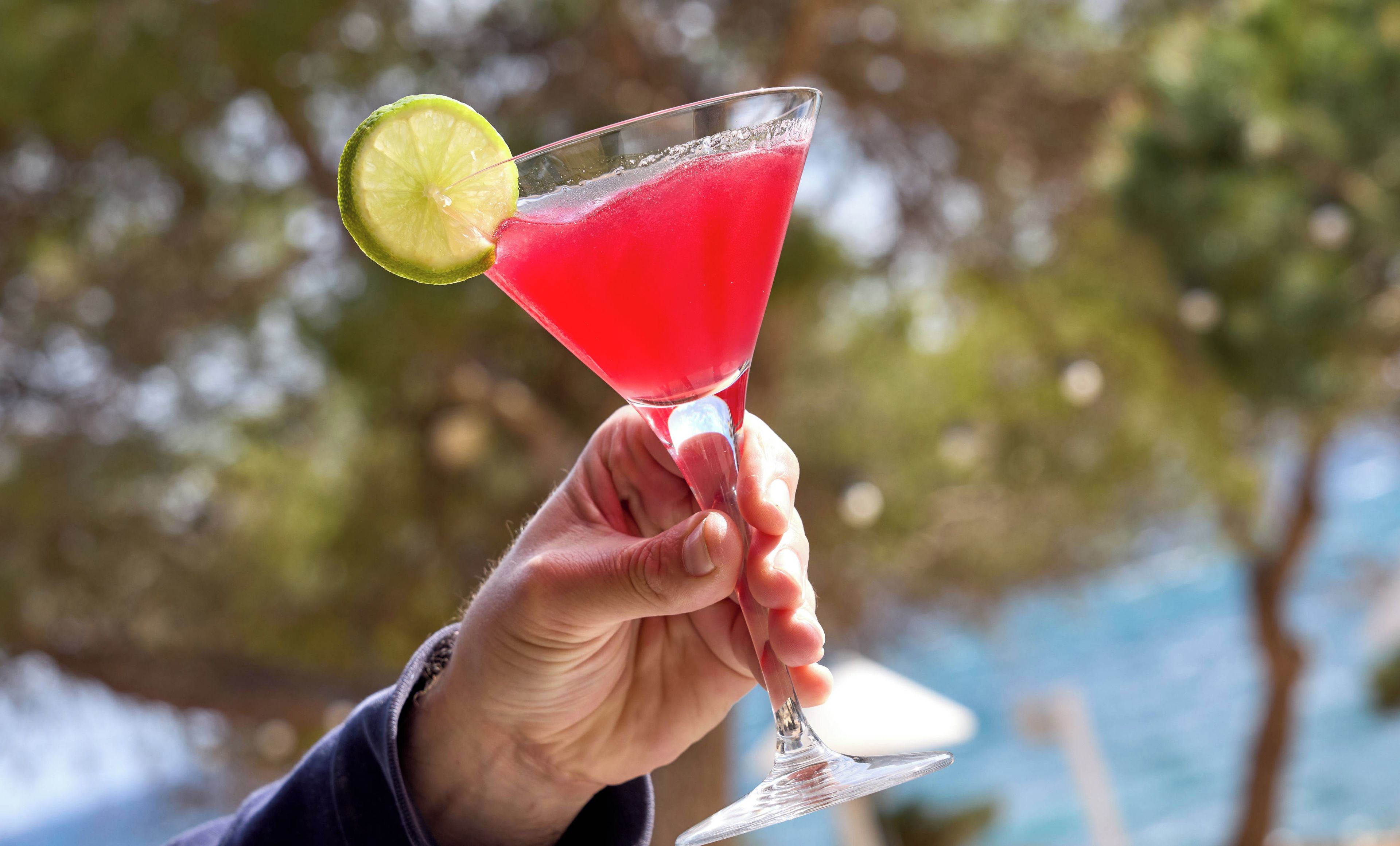 A hand holding a cocktail glass with a lime garnish, set against a blurred outdoor background.