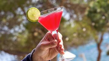 A hand holding a cocktail glass with a lime garnish, set against a blurred outdoor background.