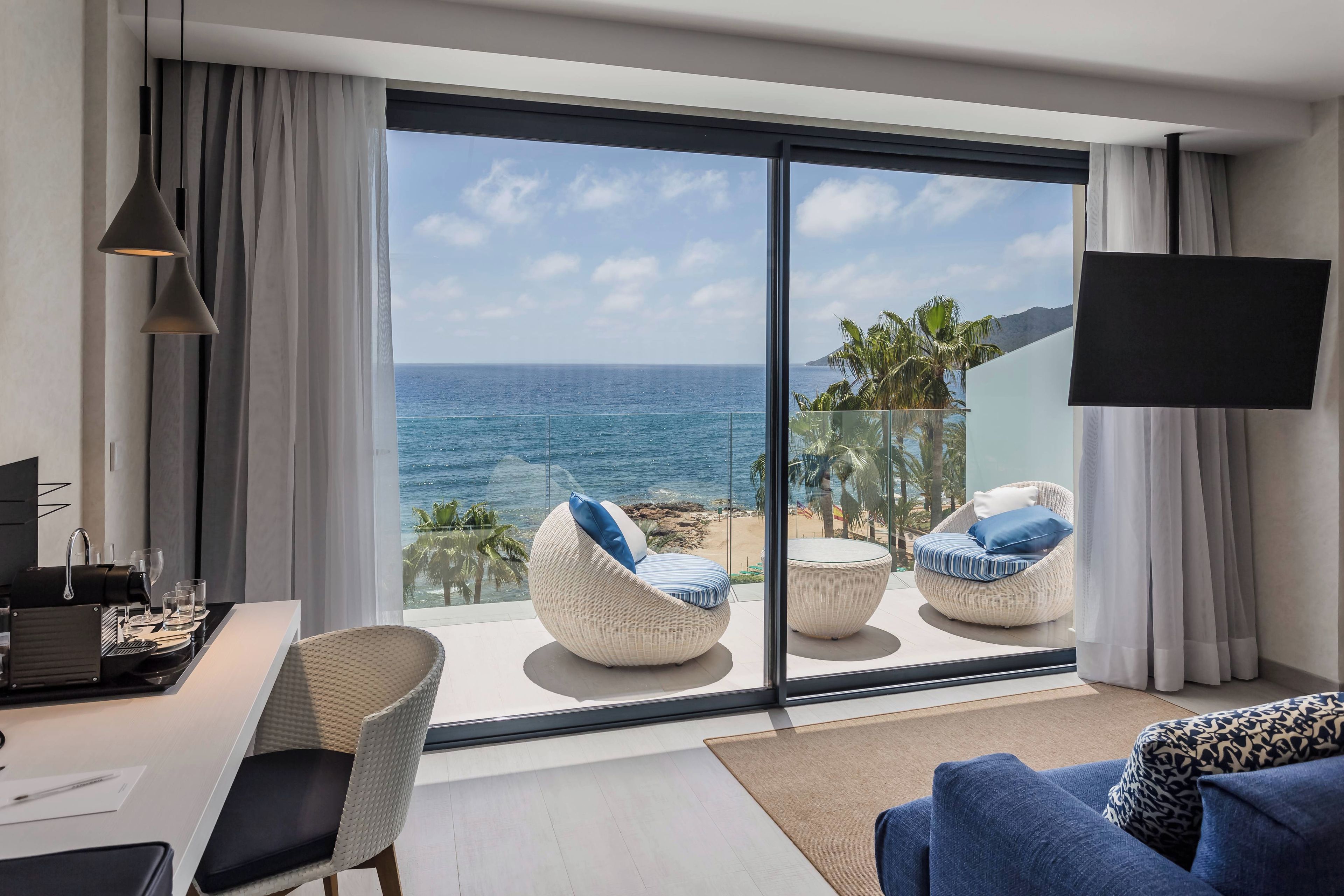 Une chambre moderne dotée d’une grande porte-fenêtre coulissante qui donne sur un balcon offrant une vue sur l’océan. On y trouve deux fauteuils en osier blancs avec des coussins bleus, un canapé blanc et un téléviseur à écran plat fixé au mur.