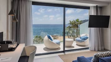 A modern room with a large sliding glass door leading to a balcony with a view of the ocean. There are two white wicker chairs with blue cushions, a white sofa, and a flat-screen TV mounted on the wall.