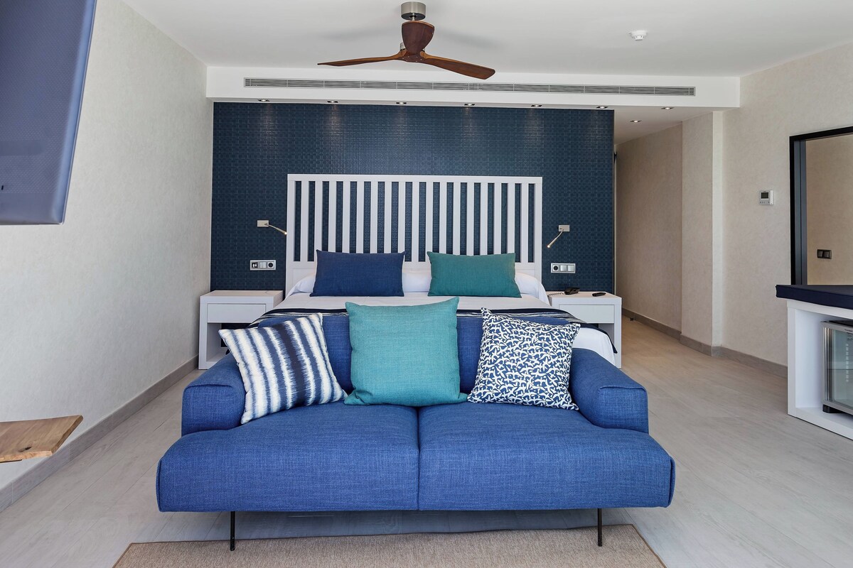 A modern bedroom with a bed, a blue sofa, a ceiling fan, and a nightstand with a lamp.