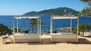 Deux espaces de détente en plein air, dotés de coussins blancs et d’une table, offrant une vue sur une étendue d’eau et un paysage montagneux.