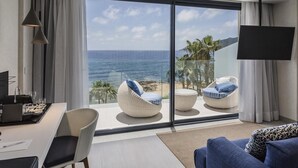 Une chambre moderne dotée d’une grande porte-fenêtre coulissante donnant sur un balcon avec vue sur l’océan. On y trouve deux fauteuils en osier blanc avec des coussins bleus, un canapé bleu et un téléviseur à écran plat fixé au mur.