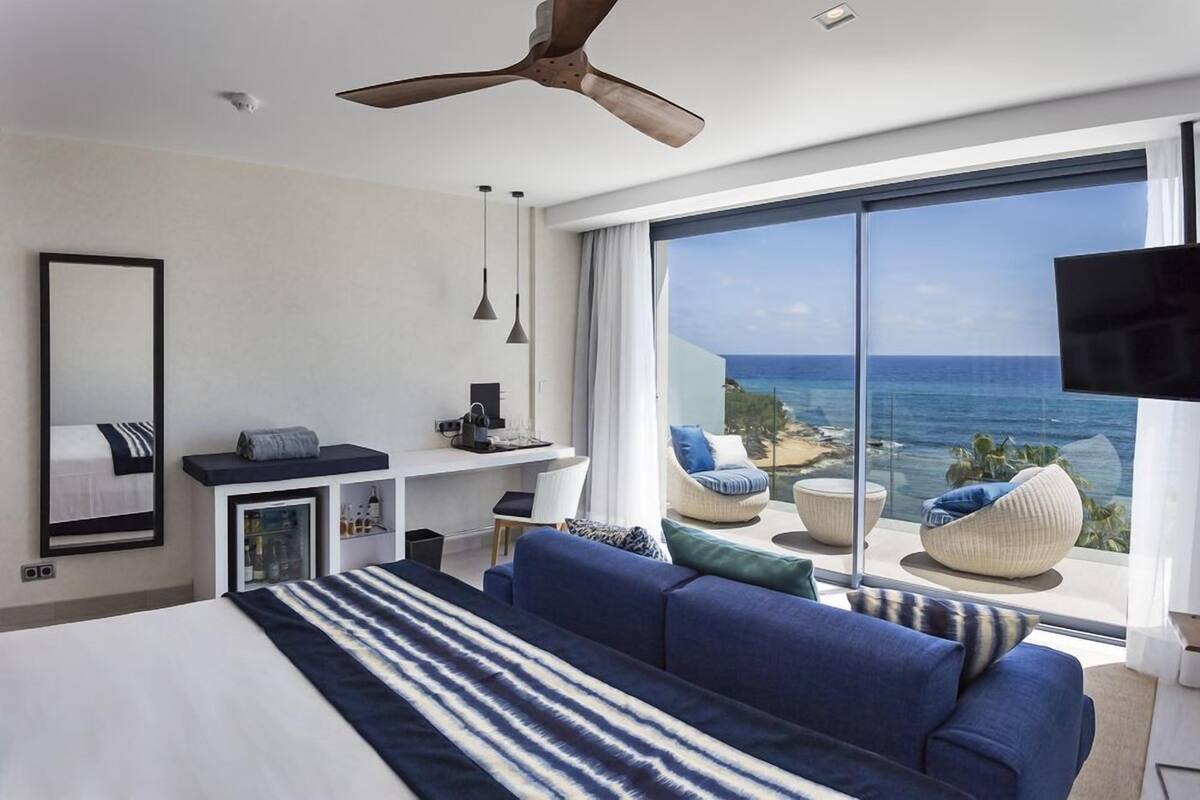 A modern hotel room with a large bed, a desk, a chair, a sofa, and a view of the ocean.