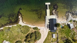 Vue aérienne d’une propriété située sur la côte, avec un quai, un grand bâtiment doté d’un toit sombre et un petit abri de jardin.
