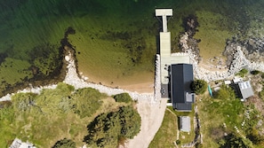 Vista aérea de una propiedad costera con un muelle, un edificio grande con techo oscuro y un cobertizo pequeño.