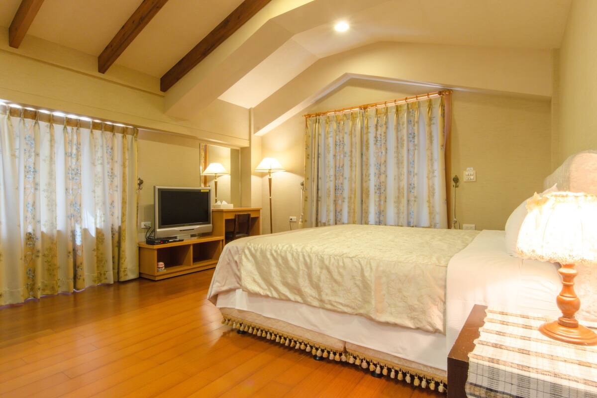 A bedroom with a large bed, wooden flooring, a television, and curtains.