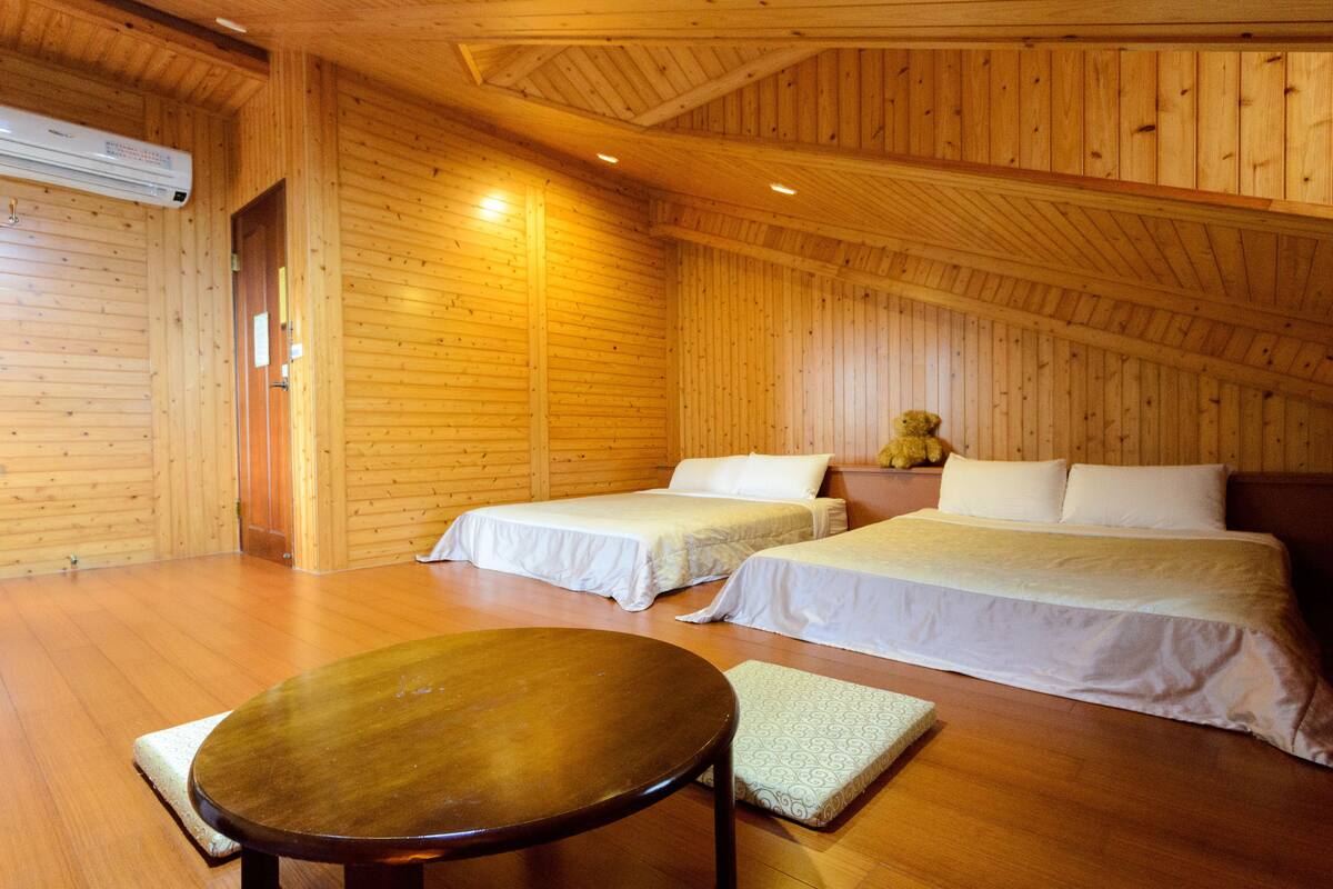 A wooden cabin room with two beds, a round table, and a teddy bear on the nightstand.
