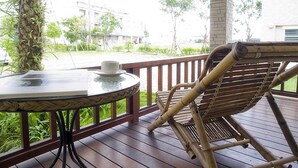Una terraza de madera con una mesa redonda y una silla de mimbre, con vista a una calle con edificios y vegetación.