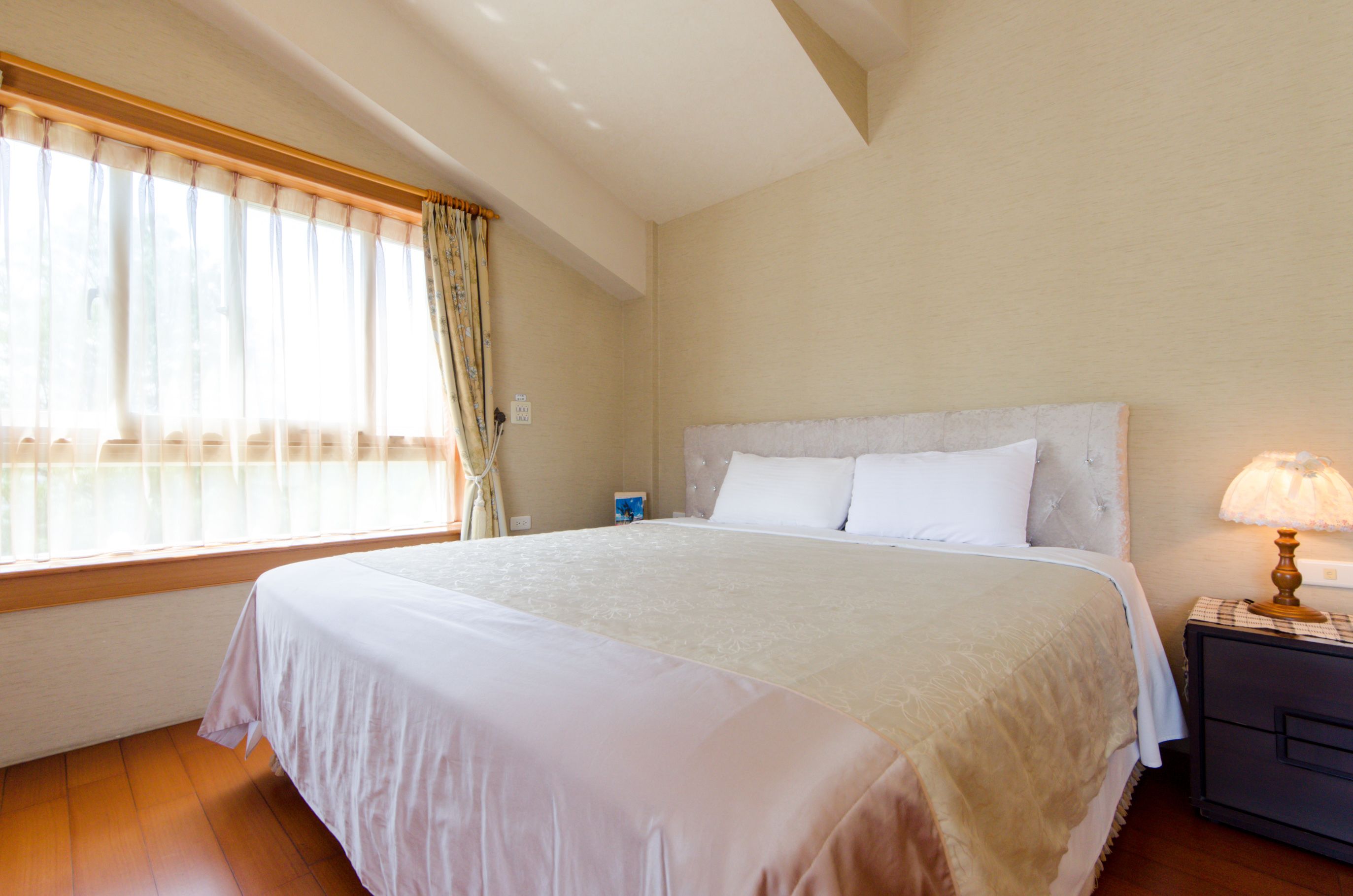 A neatly made bed with a white bedspread and pillows, a bedside table with a lamp, and a window with sheer curtains.