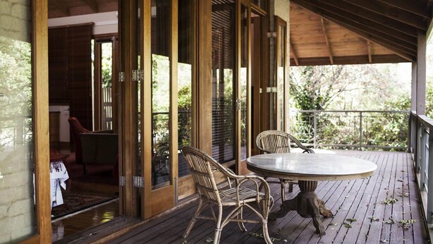 Una terraza de madera con dos sillas de mimbre y una mesa, una puerta de cristal que da al interior, y vistas a la vegetación.