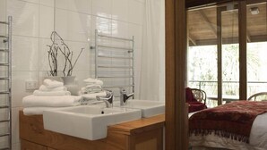 Un baño con un lavamanos grande, un espejo y un perchero para toallas. Hay un mueble de madera debajo del lavamanos y vistas a un balcón con una silla y una cama.