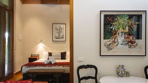 Un dormitorio con cama, mesita de noche y lámpara de pared. Se observa una obra de arte enmarcada sobre la cama y una mesa con sillas en primer plano.