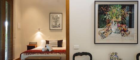 Un dormitorio con cama, mesita de noche y lámpara de pared. Se observa una obra de arte enmarcada sobre la cama y una mesa con sillas en primer plano.