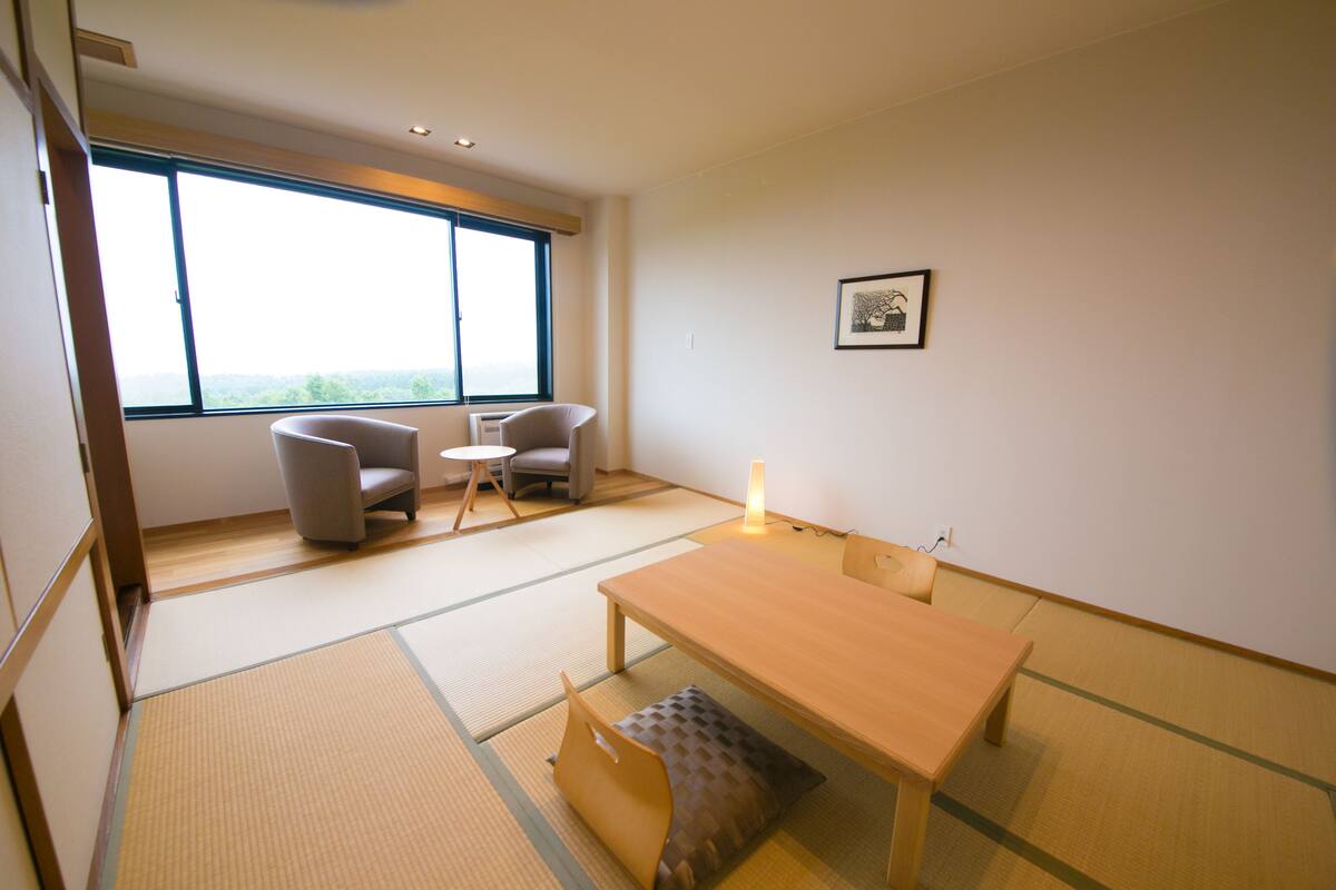 Une pièce de style japonais traditionnel, avec un sol recouvert de tatamis, une petite table basse, deux chaises et une grande fenêtre donnant sur le paysage.