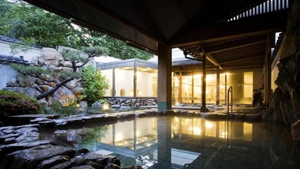 A serene indoor pool area with a stone waterfall, a wooden deck, and a traditional building with glass walls. - Dogo Onsen Hotel Tsubakikan (Matsuyama)