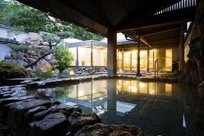 A serene indoor pool area with a stone waterfall, a wooden deck, and a traditional building with glass walls. - Dogo Onsen Hotel Tsubakikan (Matsuyama)