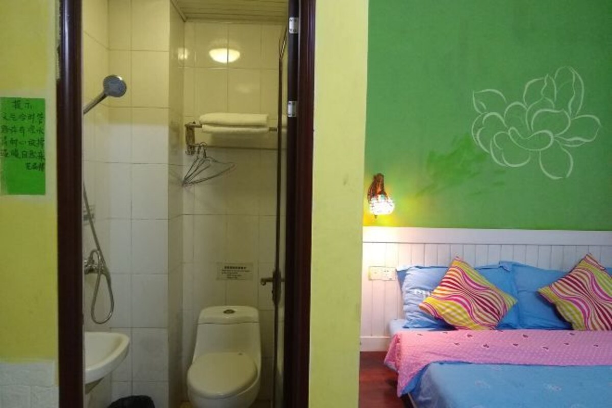 A bathroom with a toilet, sink, and shower. There is a bed with colorful pillows and a green wall with a floral design.