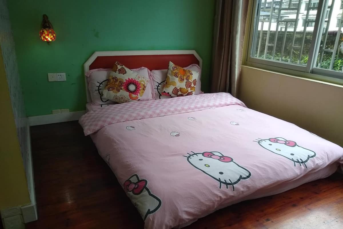 A bedroom with a pink bedspread featuring Hello Kitty, a wooden floor, a green wall, and a window with curtains.