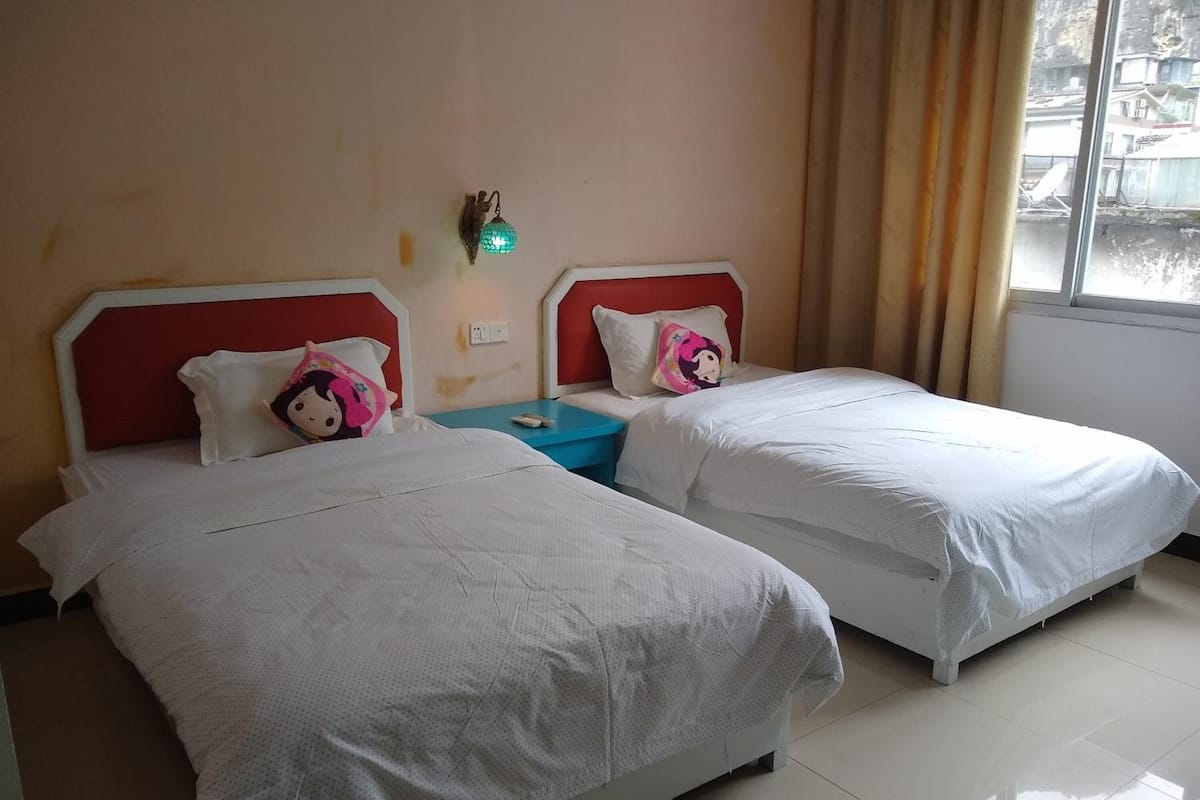 A hotel room with two single beds, a nightstand, a wall-mounted lamp, and a window with curtains.