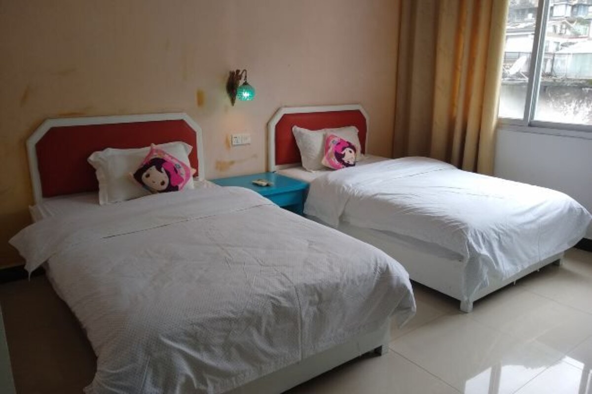 A hotel room with two beds, each with white bedding and red headboards. There is a small table between the beds with a blue tablecloth. A green wall-mounted light is visible on the wall.