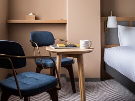 A hotel room with a bed, a desk with a chair, a lamp, and a shelf with a tissue box.