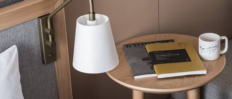 Un lit bien fait, avec du linge de lit blanc, une lampe de chevet, une petite table en bois sur laquelle se trouvent un livre et une tasse, et une prise de courant au mur.