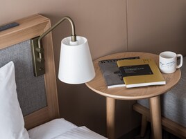 Cama bem arrumada com lençóis brancos, luminária de cabeceira, mesinha de madeira com um livro e uma caneca, e tomada na parede.