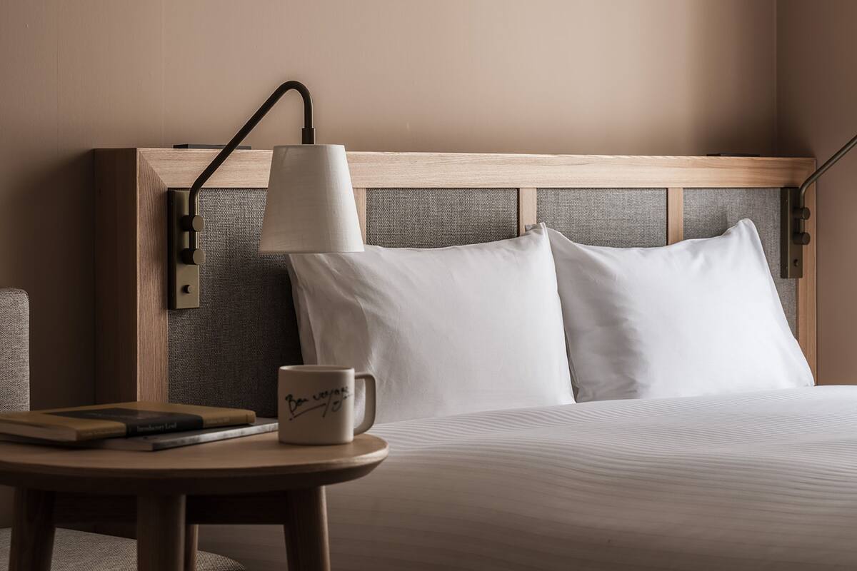 A neatly made bed with white linens, a bedside table with a lamp, and a mug with a signature.