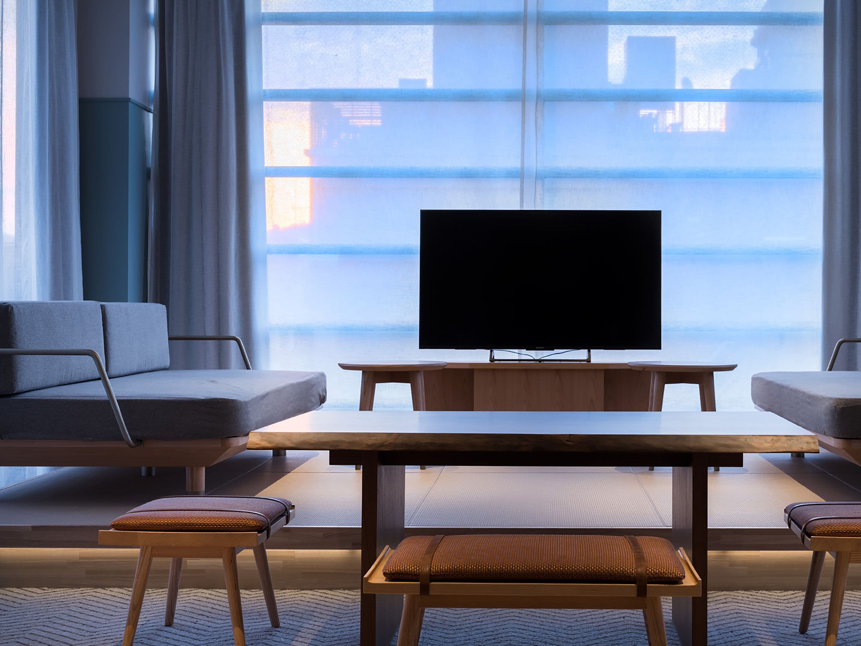 Een moderne hotelkamer met een groot raam, een flatscreen televisie, een bank, een houten tafel en twee comfortabele stoelen.
