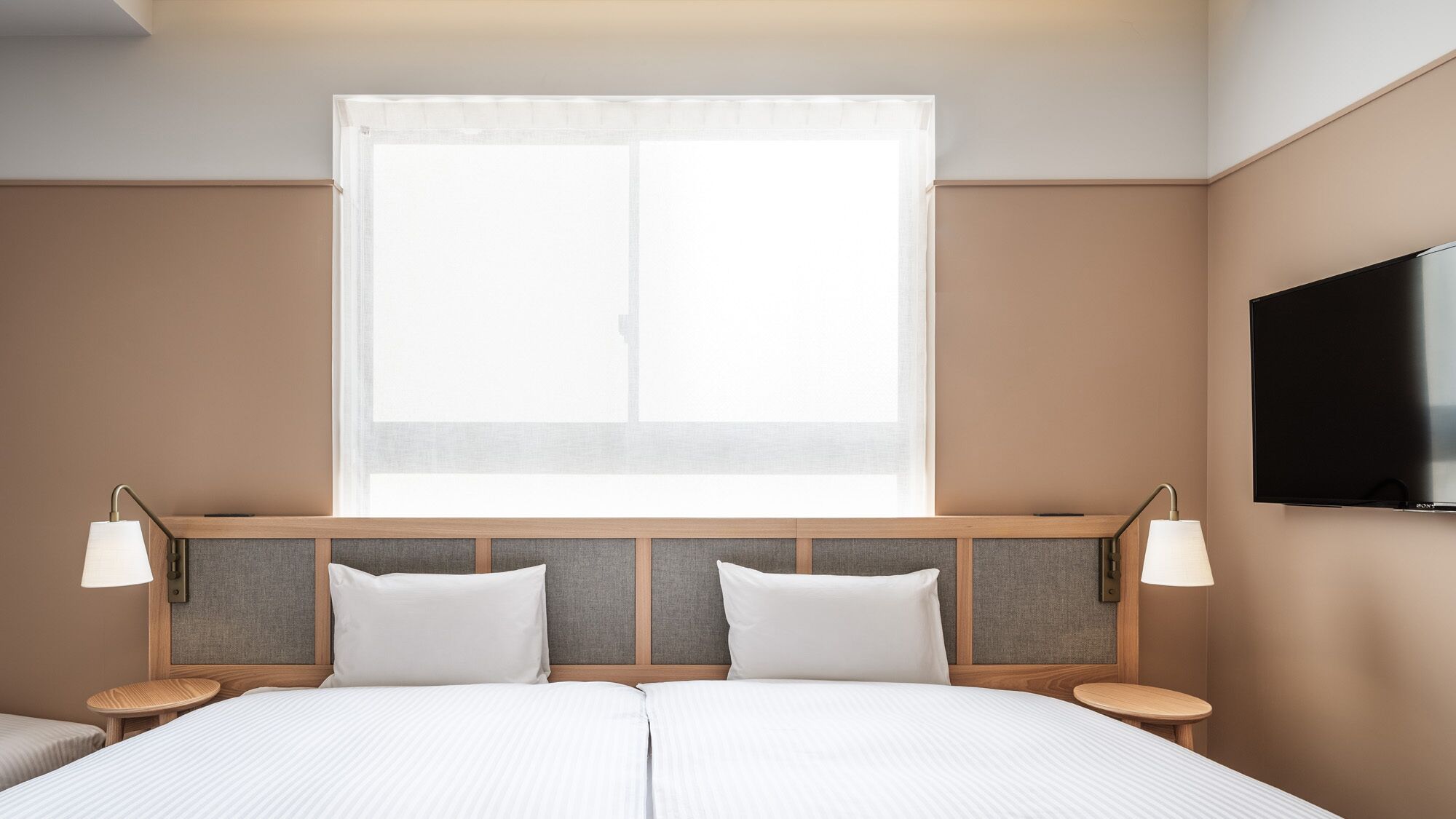 Quarto de hotel com cama grande, duas mesas de cabeceira, televisão fixada na parede e janela com cortinas translúcidas.