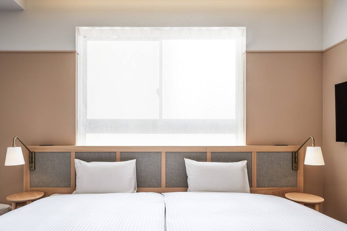 A hotel room with a large bed, two bedside tables, a mounted TV, and a window with sheer curtains.