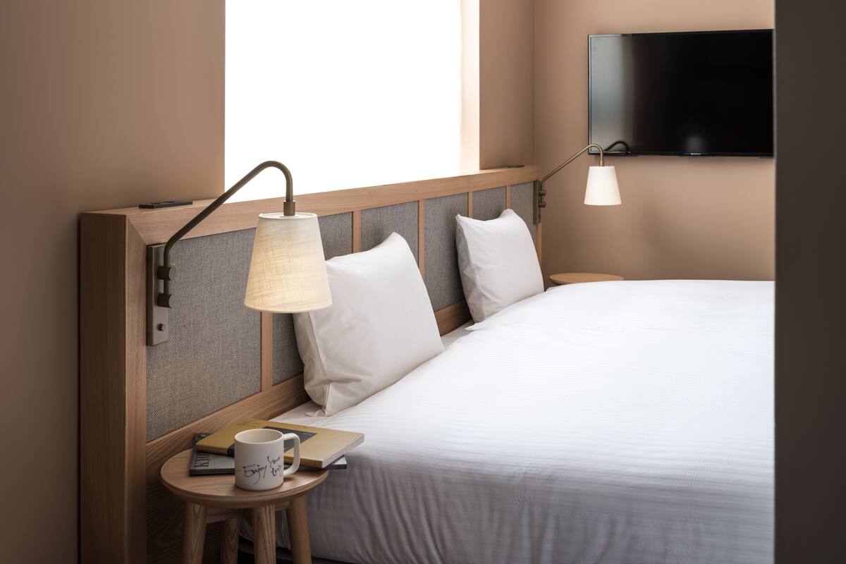 A neatly made bed with a headboard, a bedside table with a lamp, a cup, and a book. A flat-screen TV is mounted on the wall above the bed.