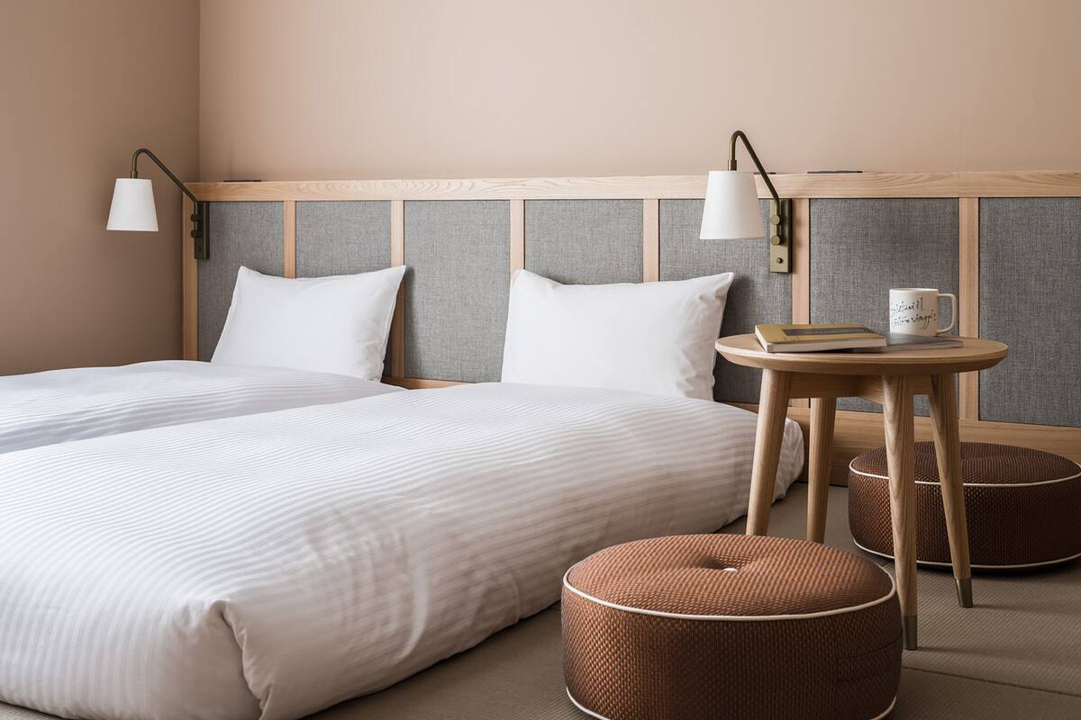 A hotel room with a large bed, two bedside lamps, a small round table with a book and a mug, and two round ottomans.