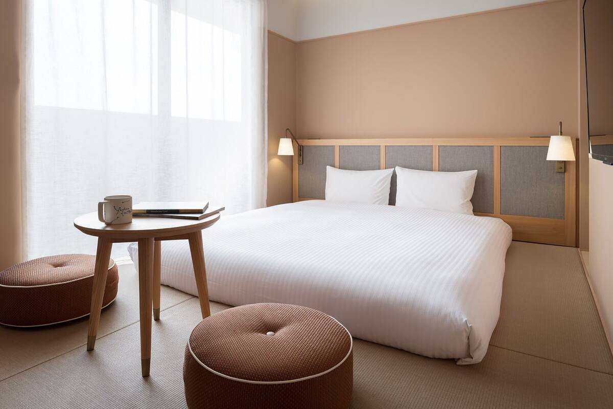 A hotel room with a large bed, a bedside table with a lamp, a small round ottoman, and a window with sheer curtains.