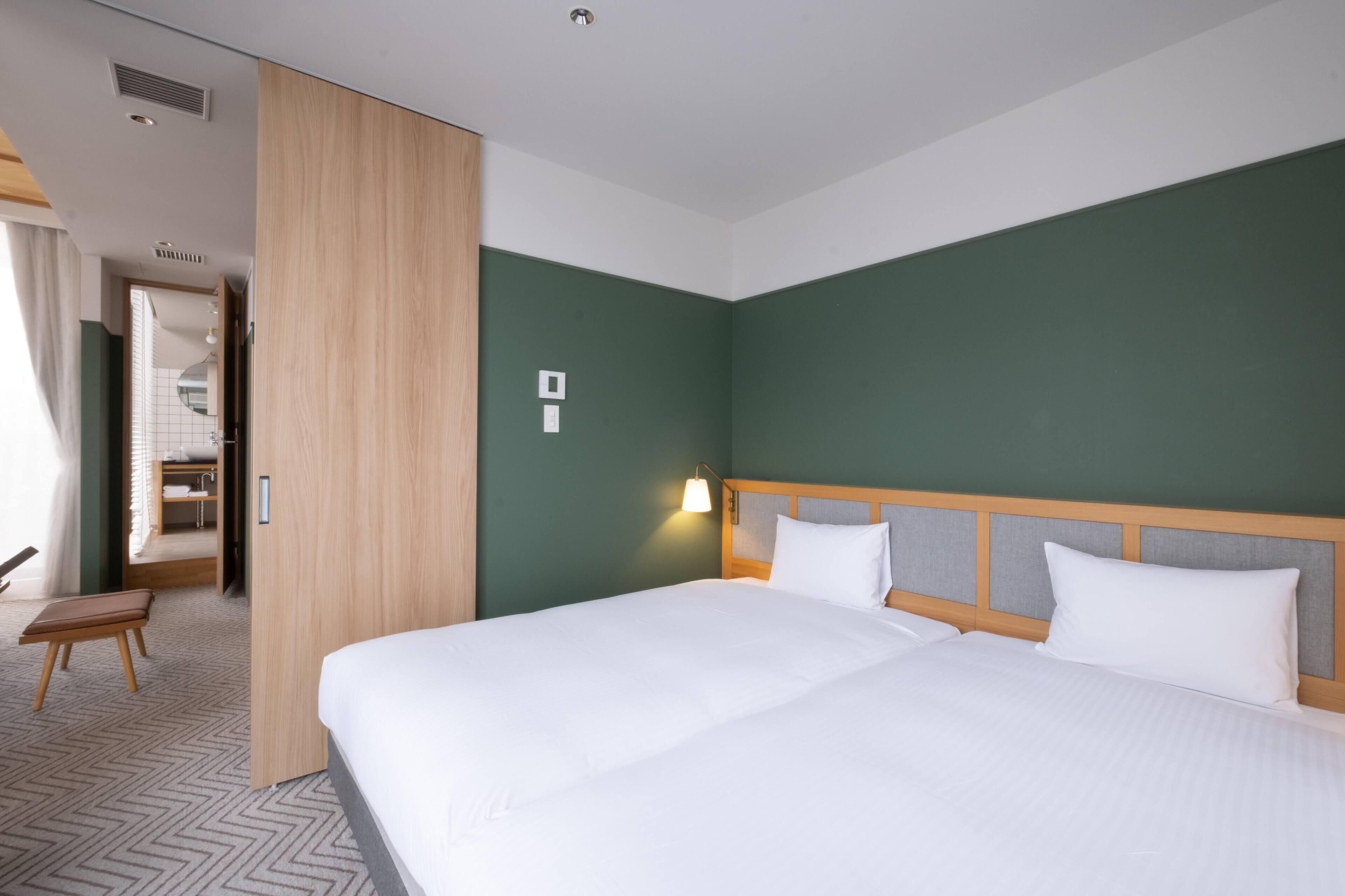 Une chambre d’hôtel avec un grand lit, une armoire en bois, un banc et une salle de bain visible à travers une porte ouverte.